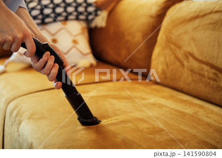 Close up of a female hands with a suction brush vacuuming the upholstery of a sofa. A woman cleans a couch using a vacuum cleaner and a nozzle for upholstered furniture. Close up of a female hands with a suction brush vacuuming the upholstery of a sofa. A woman cleans a couch using a vacuum cleaner and a nozzle for upholstered furniture. 141590804