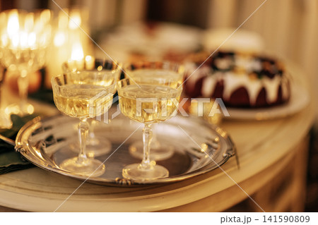 Close-up of four glasses of champagne on a beautiful metal tray. A festive cake, lighted candles and goblets of sparkling wine on the table. Close-up of four glasses of champagne on a beautiful metal tray. A festive cake, lighted candles and goblets of sparkling wine on the table. 141590809