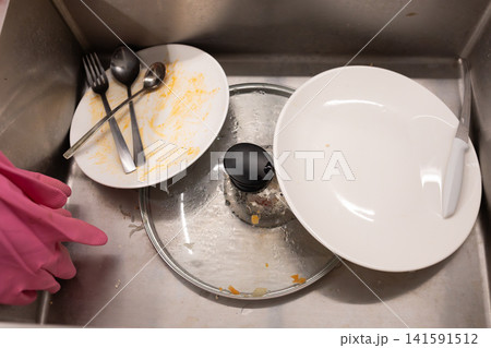 Top view of dirty dishes, plates, cutlery and a glass piled inside a stainless steel kitchen sink, showing washing up, household chores and everyday kitchen cleaning routine daily 141591512
