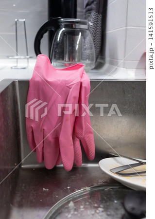 Pink rubber cleaning gloves hanging over a stainless steel kitchen sink beside dishes, showing dishwashing, household chores, hygiene and everyday domestic cleaning routine tasks 141591513