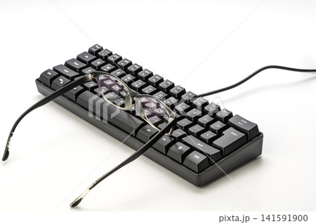 Prescription Eyeglasses With Clear Frames Resting on Compact Black Mechanical Gaming Keyboard Isolated on White Background Symbolizing Myopia and Computer Work Concept for Nearsighted Users 141591900