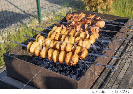 Delicious meat shashlik skewers and golden potatoes roasting on metal skewers over hot charcoal in a grill outdoors. Summer barbecue party food cooking process close-up photography. 141592287