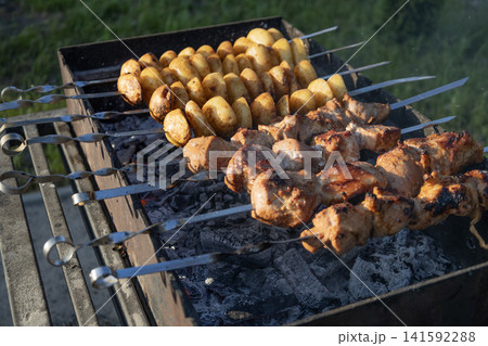 Delicious meat shashlik skewers and golden potatoes roasting on metal skewers over hot charcoal in a grill outdoors. Summer barbecue party food cooking process close-up photography. 141592288