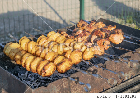 Delicious meat shashlik skewers and golden potatoes roasting on metal skewers over hot charcoal in a grill outdoors. Summer barbecue party food cooking process close-up photography. Delicious meat shashlik skewers and golden potatoes roasting on metal skewers over hot charcoal in a grill outdoors. Summer barbecue party food cooking process close-up photography. 141592293