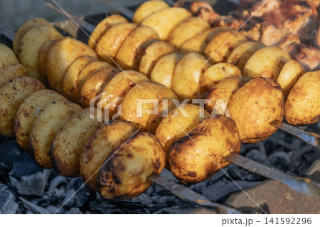 Delicious meat shashlik skewers and golden potatoes roasting on metal skewers over hot charcoal in a grill outdoors. Summer barbecue party food cooking process close-up photography. 141592296