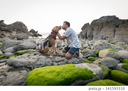 Man kneels by the water and shares a moment with his dog on rocky shore during sunset 141593310