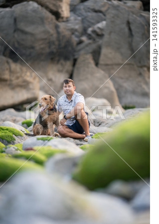 Man and dog sit on rocky shore with green moss during late afternoon Man and dog sit on rocky shore with green moss during late afternoon 141593325