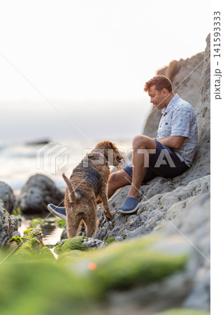 Man sits on rocks by the sea with dog during sunset near the shore Man sits on rocks by the sea with dog during sunset near the shore 141593333