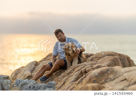 Man sits on rocks by the water with a dog during sunset near the coast 141593460
