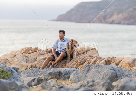 Man sits on rock by the sea with dog during sunset near coastal area Man sits on rock by the sea with dog during sunset near coastal area 141593463
