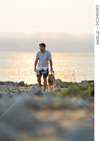 Man walks a dog along the rocky shore during sunset near the water's edge Man walks a dog along the rocky shore during sunset near the water's edge 141593465