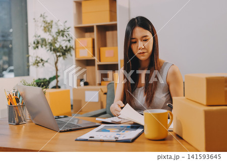 Startup SME small business entrepreneur of freelance Asian woman wearing apron using laptop and box to receive and review orders online to prepare to pack sell to customers, online sme business ideas. Startup SME small business entrepreneur of freelance Asian woman wearing apron using laptop and box to receive and review orders online to prepare to pack sell to customers, online sme business ideas. 141593645