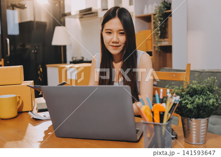 Startup SME small business entrepreneur of freelance Asian woman wearing apron using laptop and box to receive and review orders online to prepare to pack sell to customers, online sme business ideas. Startup SME small business entrepreneur of freelance Asian woman wearing apron using laptop and box to receive and review orders online to prepare to pack sell to customers, online sme business ideas. 141593647