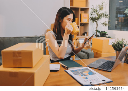 Startup SME small business entrepreneur of freelance Asian woman wearing apron using laptop and box to receive and review orders online to prepare to pack sell to customers, online sme business ideas. Startup SME small business entrepreneur of freelance Asian woman wearing apron using laptop and box to receive and review orders online to prepare to pack sell to customers, online sme business ideas. 141593654