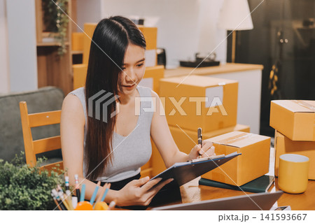 Startup SME small business entrepreneur of freelance Asian woman wearing apron using laptop and box to receive and review orders online to prepare to pack sell to customers, online sme business ideas. Startup SME small business entrepreneur of freelance Asian woman wearing apron using laptop and box to receive and review orders online to prepare to pack sell to customers, online sme business ideas. 141593667