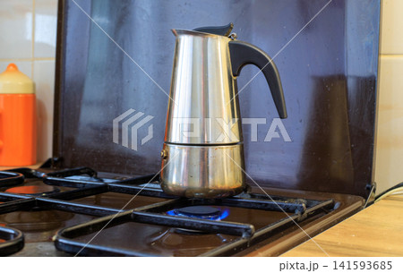 Making coffee in a moka pot. Stainless steel moka pot sits on a lit gas stove with a blue flame. Traditional espresso in a moka pot is brewed in the kitchen with orange mugs Making coffee in a moka pot. Stainless steel moka pot sits on a lit gas stove with a blue flame. Traditional espresso in a moka pot is brewed in the kitchen with orange mugs 141593685
