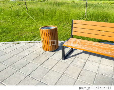 Wooden bench with metal frame positioned beside a cylindrical trash can on a paved area, surrounded by lush green grass in an urban park, providing a clean and organized outdoor space Wooden bench with metal frame positioned beside a cylindrical trash can on a paved area, surrounded by lush green grass in an urban park, providing a clean and organized outdoor space 141593811