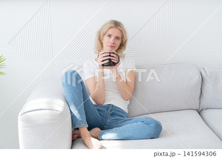 Woman enjoying cozy afternoon. Relaxed lady with coffee relaxes. Serene woman on sofa with coffee. Casual woman relaxing indoors with warm beverage and peaceful ambiance Woman enjoying cozy afternoon. Relaxed lady with coffee relaxes. Serene woman on sofa with coffee. Casual woman relaxing indoors with warm beverage and peaceful ambiance 141594306