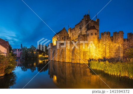 Gravensteen, a medieval castle in the city of Ghent in Belgium, Europe. Historic Stone Fortress Illuminated at Twilight, Reflected in Calm Moat Water Under a Deep Blue Sky Gravensteen, a medieval castle in the city of Ghent in Belgium, Europe. Historic Stone Fortress Illuminated at Twilight, Reflected in Calm Moat Water Under a Deep Blue Sky 141594372