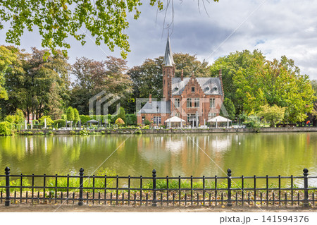 Bruges town in Belgium, Europe. Minnewater Castle, A picturesque historic building with a steeple and gabled roof, nestled beside a beautiful lake on a cloudy day, reflected in the water Bruges town in Belgium, Europe. Minnewater Castle, A picturesque historic building with a steeple and gabled roof, nestled beside a beautiful lake on a cloudy day, reflected in the water 141594376