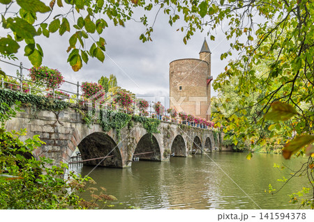 Bruges town in Belgium, Europe. Gunpowder Tower Poertoren with Ancient Stone Arch Bridge Spanning a Calm River, Adorned with Lush Green Ivy and Colorful Flowers, Under a Cloudy Sky Bruges town in Belgium, Europe. Gunpowder Tower Poertoren with Ancient Stone Arch Bridge Spanning a Calm River, Adorned with Lush Green Ivy and Colorful Flowers, Under a Cloudy Sky 141594378