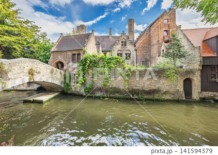 Bruges town in Belgium, Europe. Historic stone bridge arches over a canal beside old brick buildings adorned with lush green ivy under a vibrant blue sky Bruges town in Belgium, Europe. Historic stone bridge arches over a canal beside old brick buildings adorned with lush green ivy under a vibrant blue sky 141594379