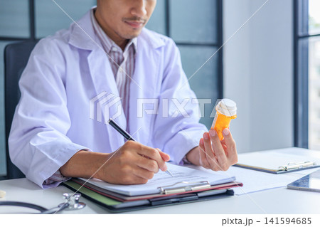 Doctor writing prescription order and holding medication bottle at desk in clinic. Doctor writing prescription order and holding medication bottle at desk in clinic. 141594685
