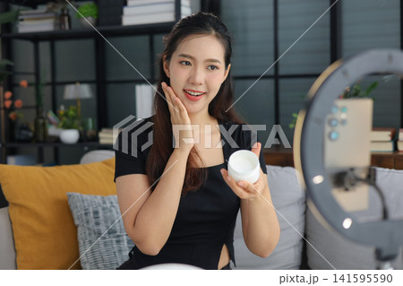 Asian woman filming beauty vlog tutorial with ring light, reviewing cosmetics cream moisturizer or lotion. 141595590