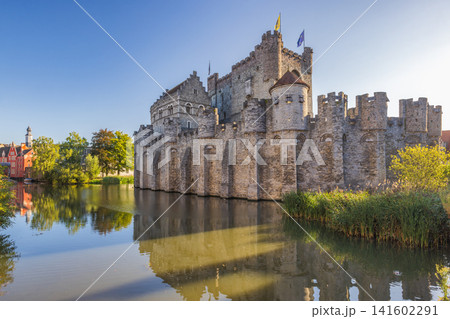 Gravensteen, a medieval castle in the city of Ghent in Belgium, Europe. Historic Stone Fortress with Turrets Reflected in Calm River Water Under a Bright Blue Sky on a Sunny Day 141602291