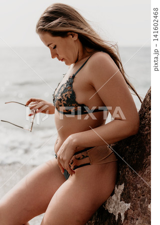 Woman beach swimwear leaning against a coastal rock while holding sunglasses during a serene summer day at the ocean Woman beach swimwear leaning against a coastal rock while holding sunglasses during a serene summer day at the ocean 141602468
