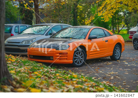 Autumn. Orange modified sports coupe with black hood parked beside other vehicles on a tree-lined street covered with fallen leaves Autumn. Orange modified sports coupe with black hood parked beside other vehicles on a tree-lined street covered with fallen leaves 141602847