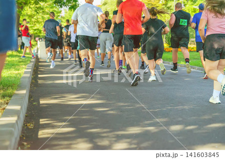 Participants run through park, Become a Hero obstacle race in Burgas. Adult participants run along a park pathway during the Become a Hero sport obstacle race. Rear view Participants run through park, Become a Hero obstacle race in Burgas. Adult participants run along a park pathway during the Become a Hero sport obstacle race. Rear view 141603845