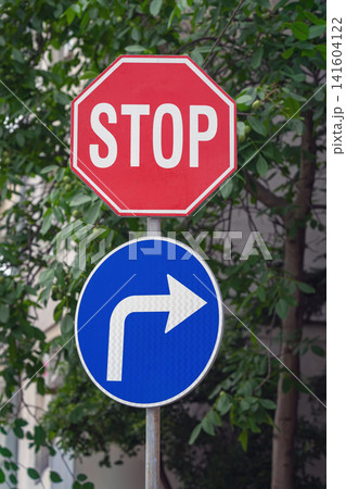 Red stop sign above a blue mandatory right turn sign against green foliage. Urban road signs representing traffic regulations, driving rules, road safety and direction 141604122