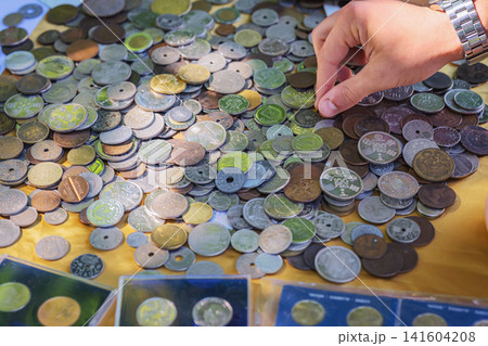 Collector Selecting Vintage Coin from Numismatic Collection. Hand selects a vintage coin from large assortment of old international currency displayed on flea market table. Numismatic scene Collector Selecting Vintage Coin from Numismatic Collection. Hand selects a vintage coin from large assortment of old international currency displayed on flea market table. Numismatic scene 141604208