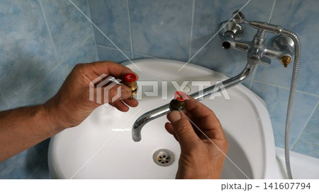 A technician holds two removed faucet valves in front of a disassembled faucet. He's assessing the condition of the parts before replacing or repairing them, above the sink in the bathroom 141607794