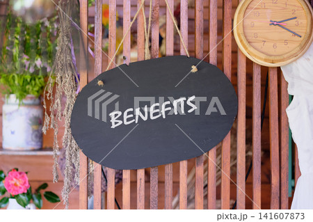 A sign hangs at a market stall showing the word benefits. The stall features wooden elements and has plants in containers nearby A sign hangs at a market stall showing the word benefits. The stall features wooden elements and has plants in containers nearby 141607873