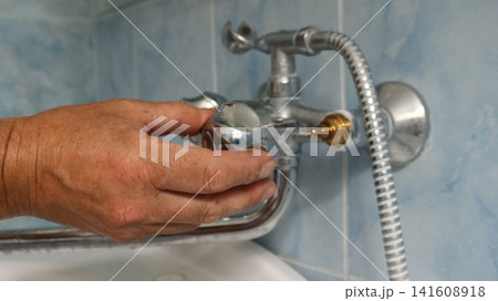 A man's hand places a chrome flywheel onto the installed valve body of a two-handle faucet. The second handle is still missing, revealing a brass component. A man's hand places a chrome flywheel onto the installed valve body of a two-handle faucet. The second handle is still missing, revealing a brass component. 141608918