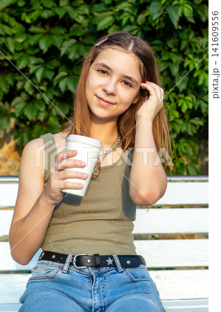 Teen girl enjoying coffee on a park bench in casual summer outfit with a gentle smile Teen girl enjoying coffee on a park bench in casual summer outfit with a gentle smile 141609556