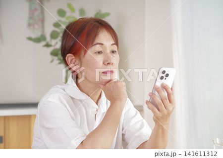スマートフォンを見ながら悩む中高年女性 スマートフォンを見ながら悩む中高年女性 141611312