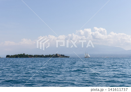 Sailboat near a palace island on Lake Garda, open blue water and distant mountains create a serene summer travel scene with wooded shoreline clouds spacious sky and lakeside vacation mood 141611737