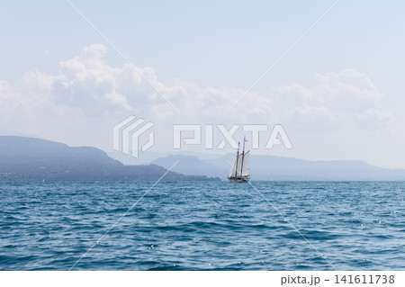 Sailboat on Lake Garda seen from the water on a summer day, blue waves stretch across the foreground while hazy mountain shores and large white clouds create an airy travel landscape with calm open Sailboat on Lake Garda seen from the water on a summer day, blue waves stretch across the foreground while hazy mountain shores and large white clouds create an airy travel landscape with calm open 141611738