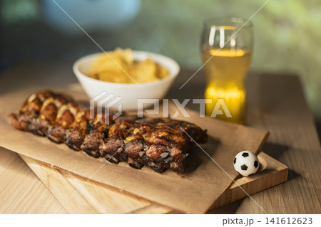 Barbecue ribs on a wooden board, beer and snacks arranged for a soccer viewing party, creating a cozy football night atmosphere. Barbecue ribs on a wooden board, beer and snacks arranged for a soccer viewing party, creating a cozy football night atmosphere. 141612623