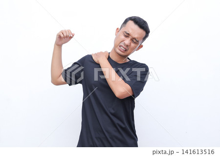 Man grips shoulder, showing pain. His face shows discomfort, eyes squeezed shut. He wears a plain black t-shirt. White background isolates the subject clearly. Ideal for health, fitness 141613516