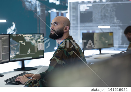 Military analyst working at command post workstation watching tracking system on wall displays. Intelligence officer tracks satellite radar surveillance to extract military target information Military analyst working at command post workstation watching tracking system on wall displays. Intelligence officer tracks satellite radar surveillance to extract military target information 141616922