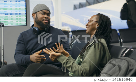 Black lovers talking about journey and waiting for flight departure, seated in airport lounge with runway view. Couple enjoying cute romantic moment, discussing leisure travel plan. Black lovers talking about journey and waiting for flight departure, seated in airport lounge with runway view. Couple enjoying cute romantic moment, discussing leisure travel plan. 141616940