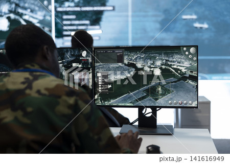 Man in military command post working on PC, looking at surveillance footage on monitor. Military operator watches intelligence feed at workstation desk, close up shot 141616949