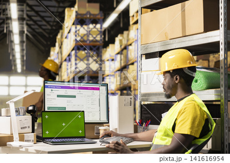 Huge distribution center worker verifying parcel details with isolated display, handling order processing in e-commerce fulfillment center. Large business retail industrial operations. Huge distribution center worker verifying parcel details with isolated display, handling order processing in e-commerce fulfillment center. Large business retail industrial operations. 141616954