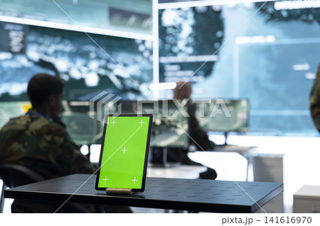 Close up of green screen tablet on military command post table in front of soldier working. Mockup device next to army personnel at workstations across operations room Close up of green screen tablet on military command post table in front of soldier working. Mockup device next to army personnel at workstations across operations room 141616970