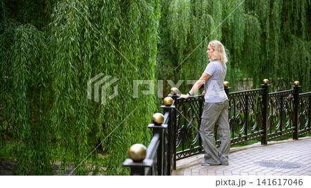 Middle-aged Caucasian woman standing by a metal fence in a park. Blonde female looking at lush green weeping willow trees. Casual outdoor lifestyle, background and leisure Middle-aged Caucasian woman standing by a metal fence in a park. Blonde female looking at lush green weeping willow trees. Casual outdoor lifestyle, background and leisure 141617046