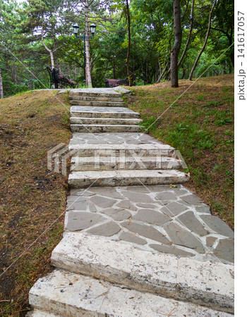 Vertical photo of stone stairs leading up a hill in a green park. Rustic pathway, staircase with vintage lamppost and wooden benches among trees Vertical photo of stone stairs leading up a hill in a green park. Rustic pathway, staircase with vintage lamppost and wooden benches among trees 141617057